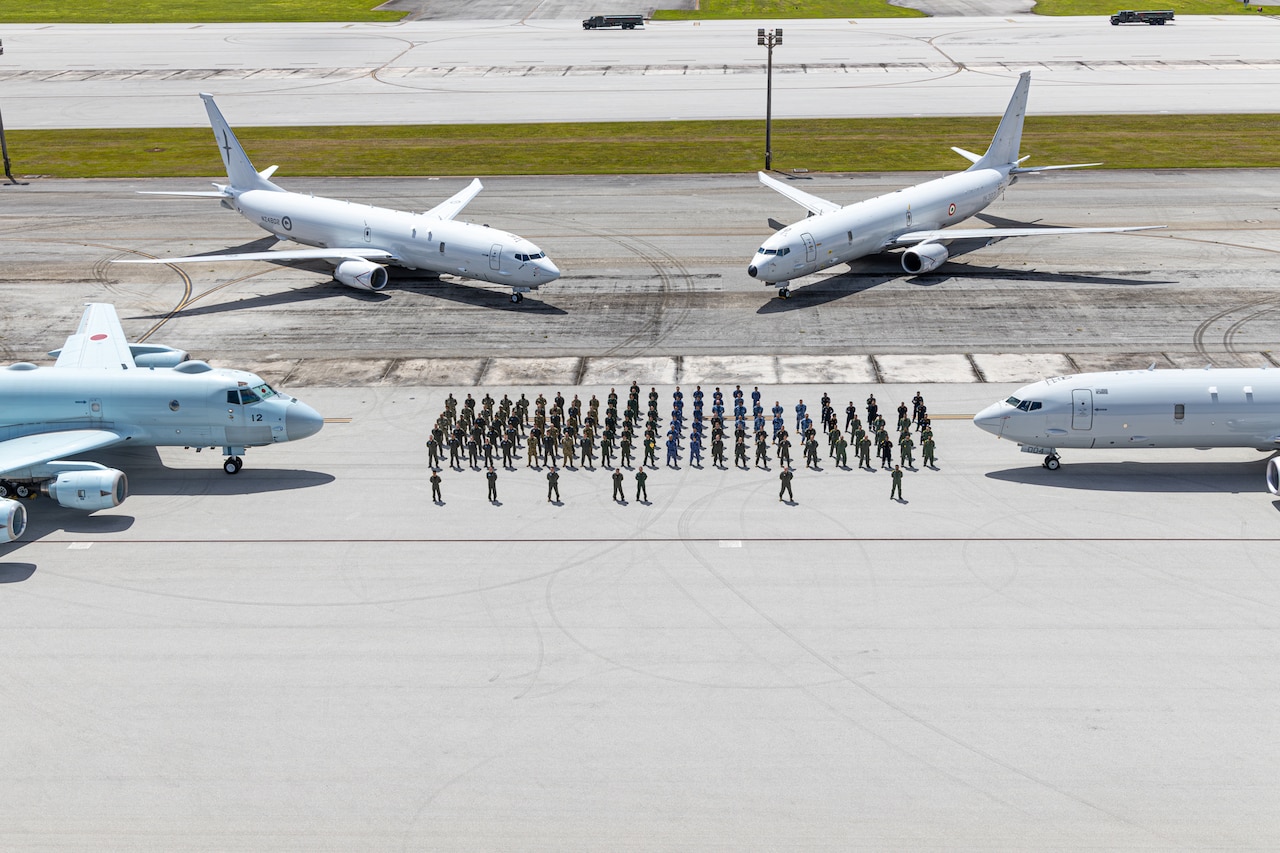 Dozens of people wearing military uniforms stand in formation on a tarmac while surrounded by four large military aircraft.