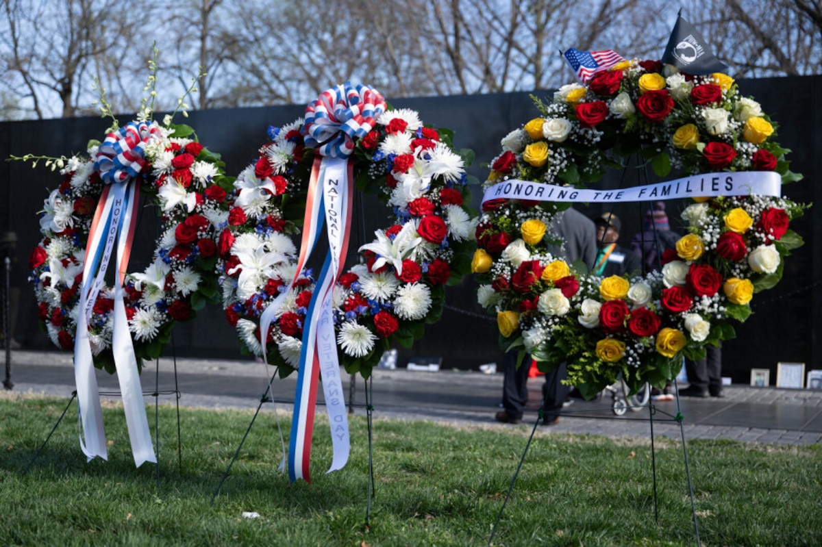 Three large, bright flower wreaths are resting on the grass outside in front of a black stone wall.