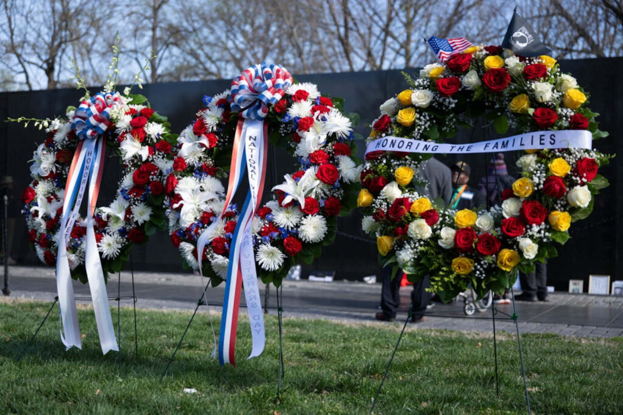 Three large, bright flower wreaths are resting on the grass outside in front of a black stone wall.