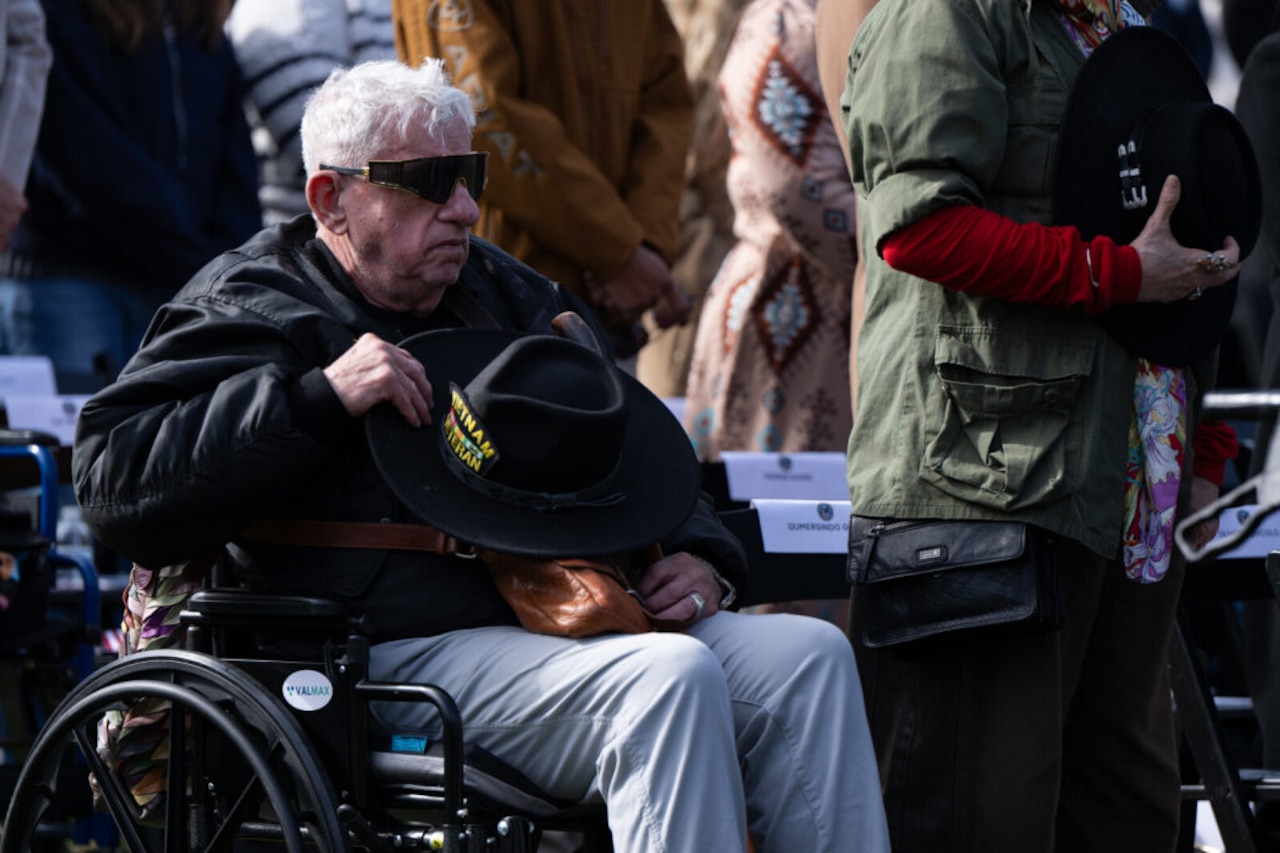 An elderly man sitting in a wheelchair, wearing sunglasses and holding a dark hat that reads "Vietnam Veteran," sits outdoors, observing what is happening during a ceremony.