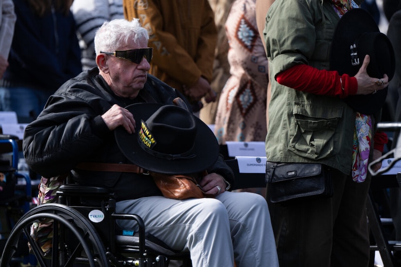 An elderly man sitting in a wheelchair, wearing sunglasses and holding a dark hat that reads "Vietnam Veteran," sits outdoors, observing what is happening during a ceremony.