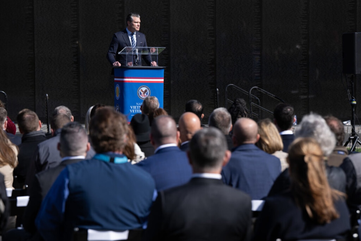A man in business attire speaks outside behind a lectern to a large group of people with their backs to the camera.