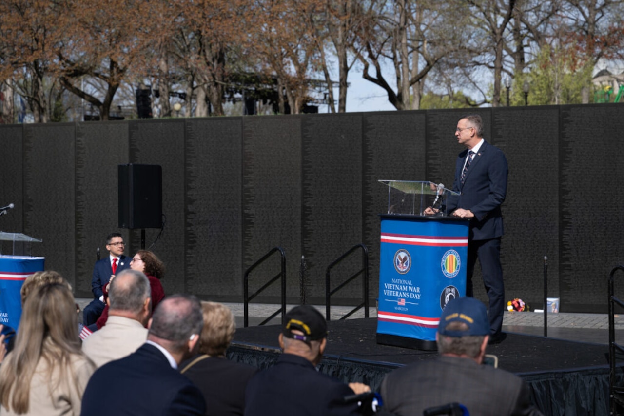 A man in business attire speaks outside behind a lectern to a group of people seated in front of him. A large black wall is behind the man.
