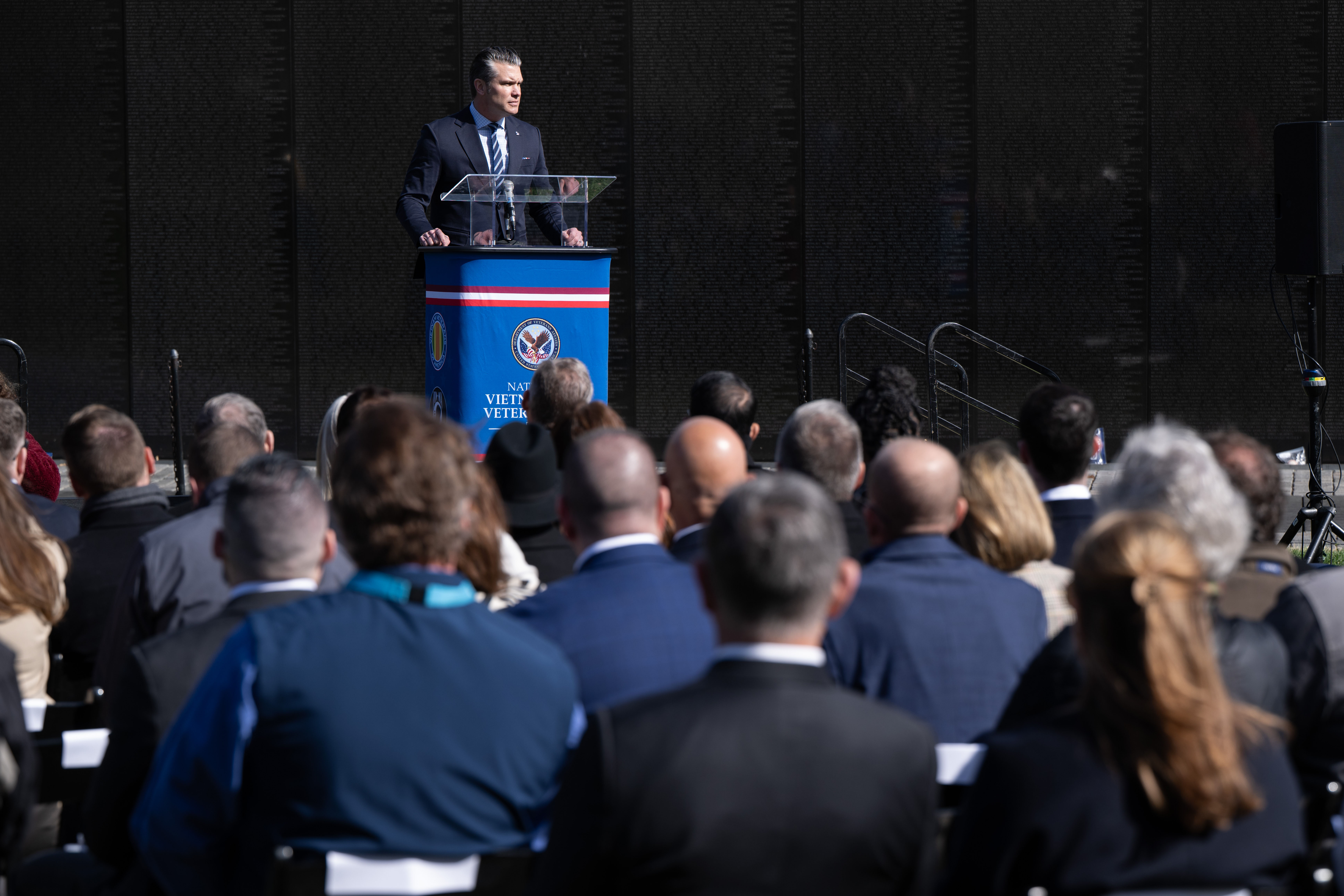 A person in a business suit stands at a lectern outdoors and speaks to a seated crowd.