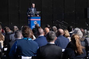 Secretary of War Pete Hegseth stands at a lectern outdoors and speaks to a seated crowd.