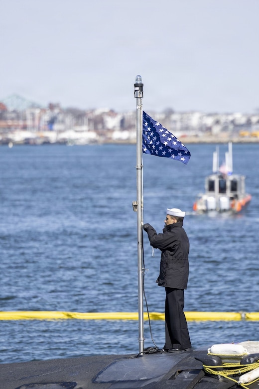 A man in a military dress uniform raises a flag while standing on top of a submarine.