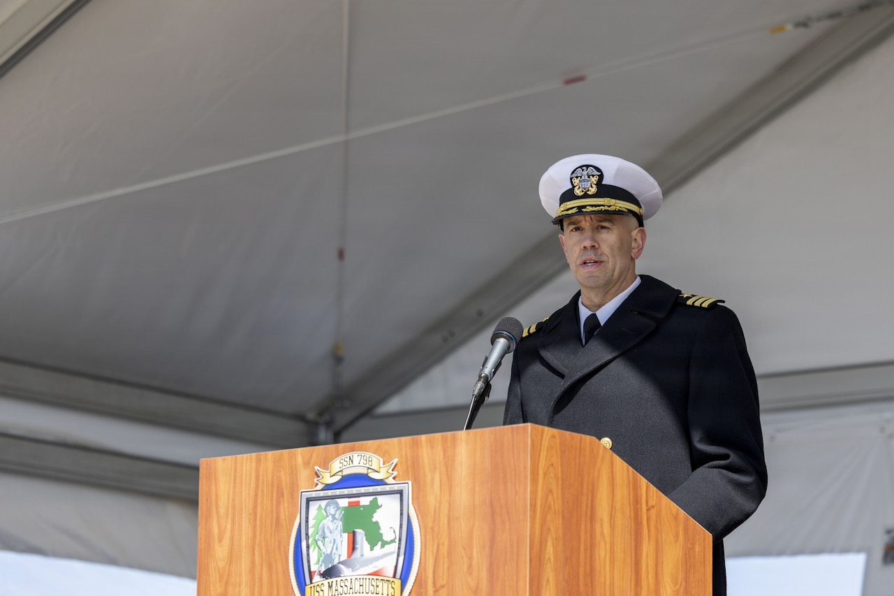 A man in a military dress uniform speaks into a microphone while standing behind a lectern.