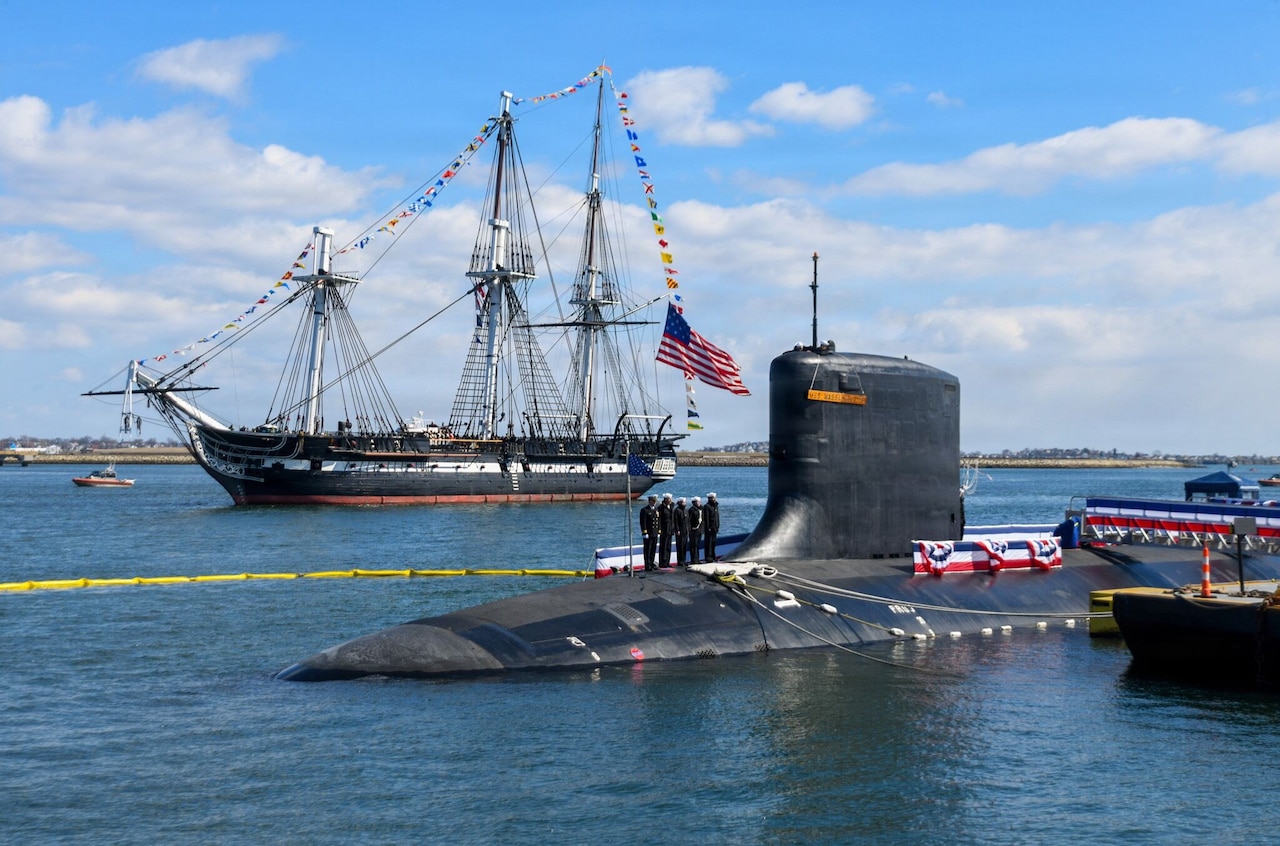 A Revolutionary War-era ship with the American flag flying from the stern sails past a submarine moored to a dock as five people in military uniforms stand on the sub.