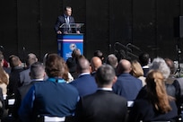 Secretary of War Pete Hegseth stands at a lectern outdoors and speaks to a seated crowd.