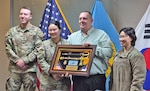 A man holds a shadow box in front of flags while posing with three soldiers.