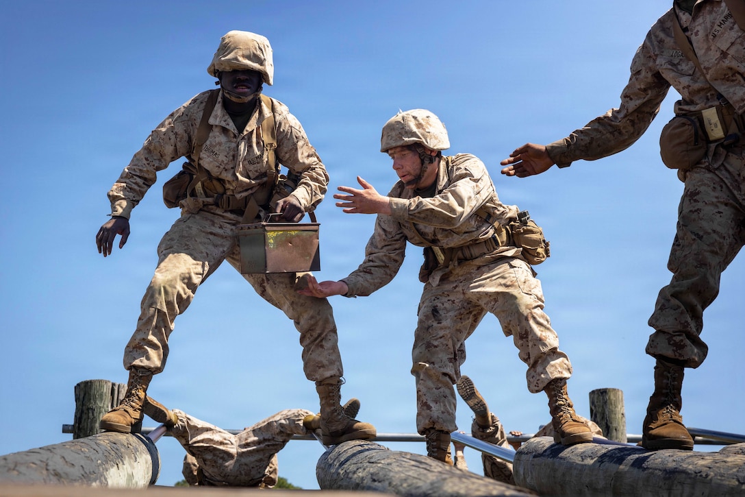Three Marine Corps recruits walk across elevated logs while passing a metal box against a blue sky.