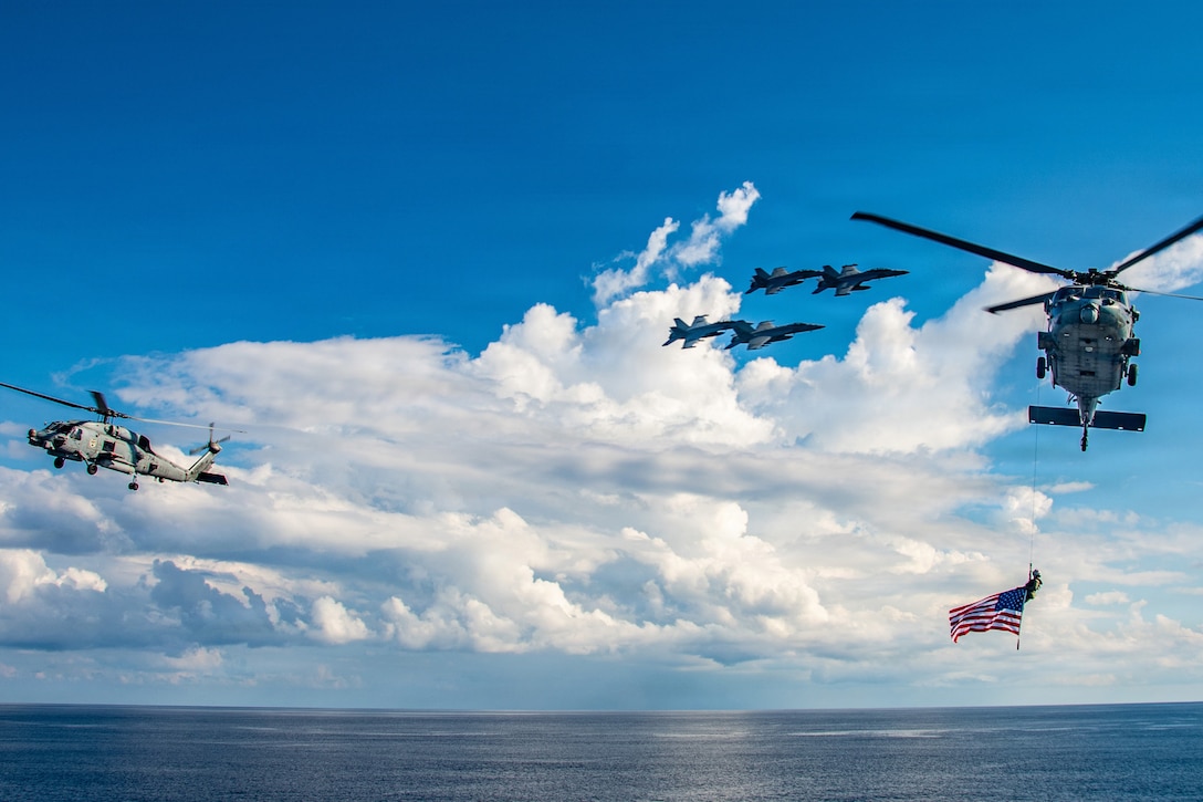 Two helicopters, one displaying an American flag, fly on opposite sides in a blue sky over a body of water as four aircraft fly in formation nearby.