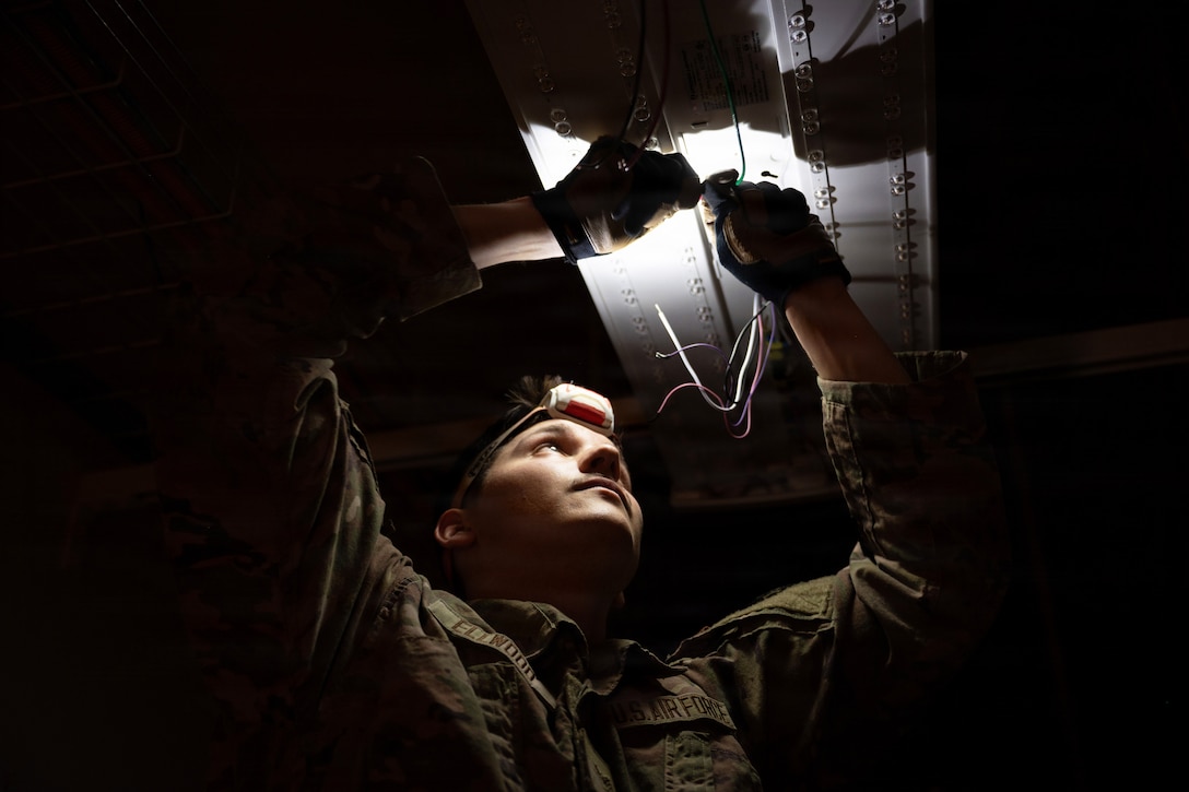 An airman works on a light fixture near hanging wires in the dark illuminated by a dim light.