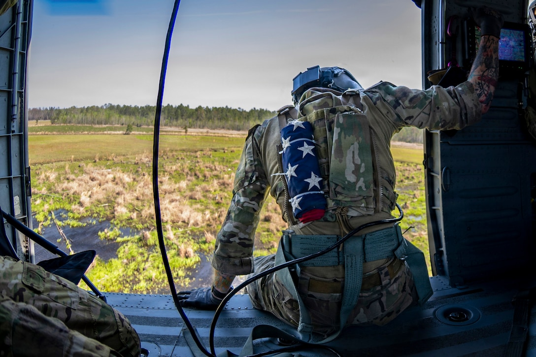An airman carrying a folded American flag on their back sits in the open doorway of an aircraft as it flies over a field during the day.