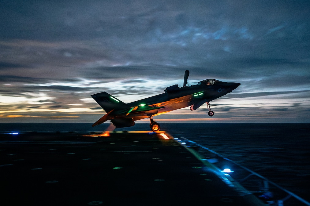 A Marine Corps aircraft takes off from a ship in the dark illuminated by a yellowish sky and blue and green lights.