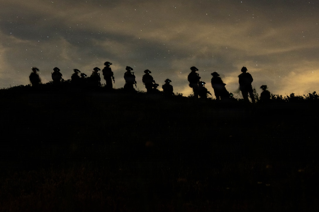 About a dozen Marines, shown in silhouette, move on hilly terrain against a dark sky.