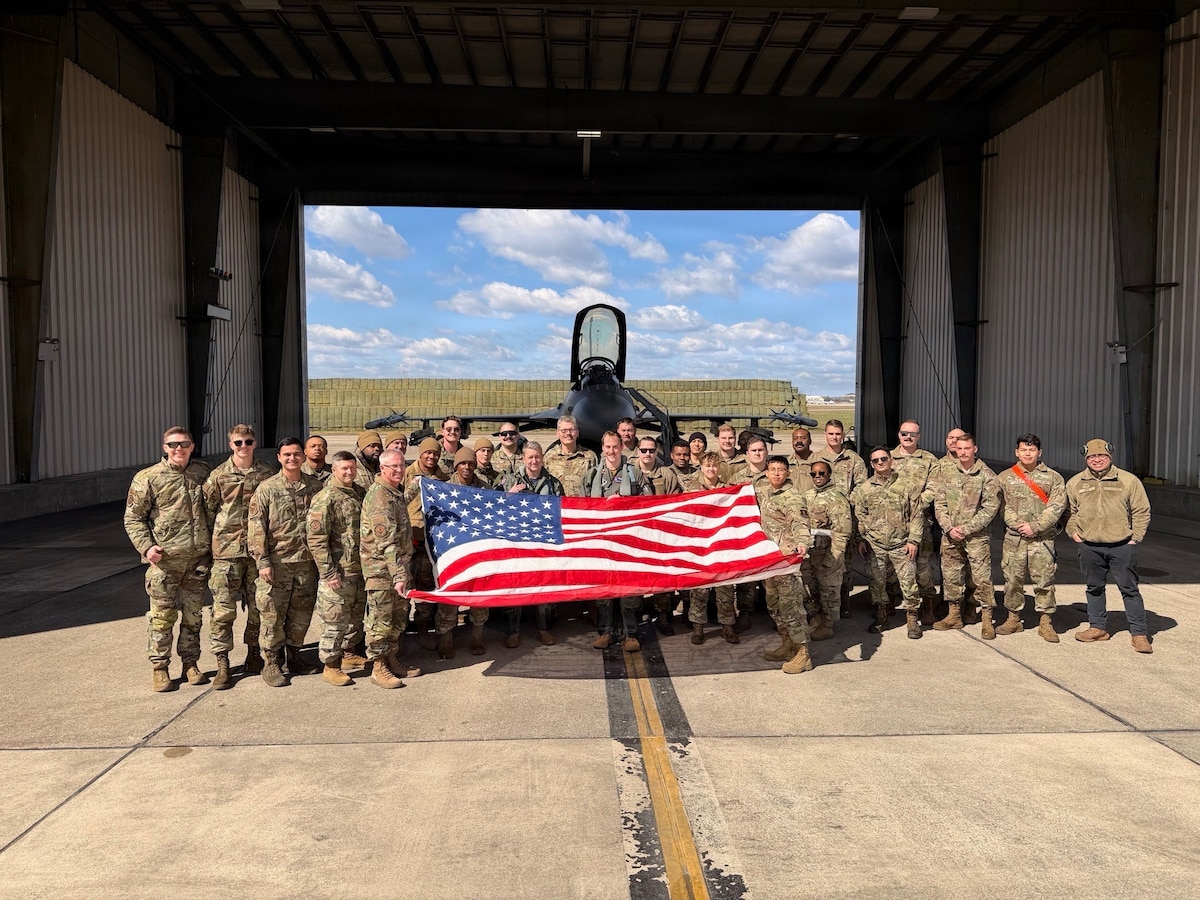 Dozens of people in military camouflage uniforms pose for a photo in front of an aircraft hangar while holding up an American flag. A military jet is in the background.