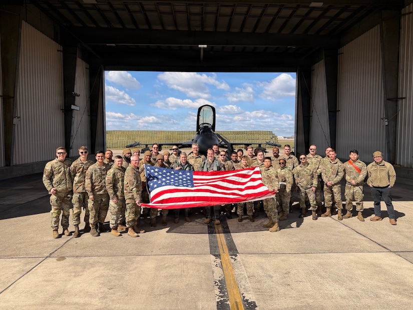 Dozens of people in military camouflage uniforms pose for a photo in front of an aircraft hangar while holding up an American flag. A military jet is in the background.