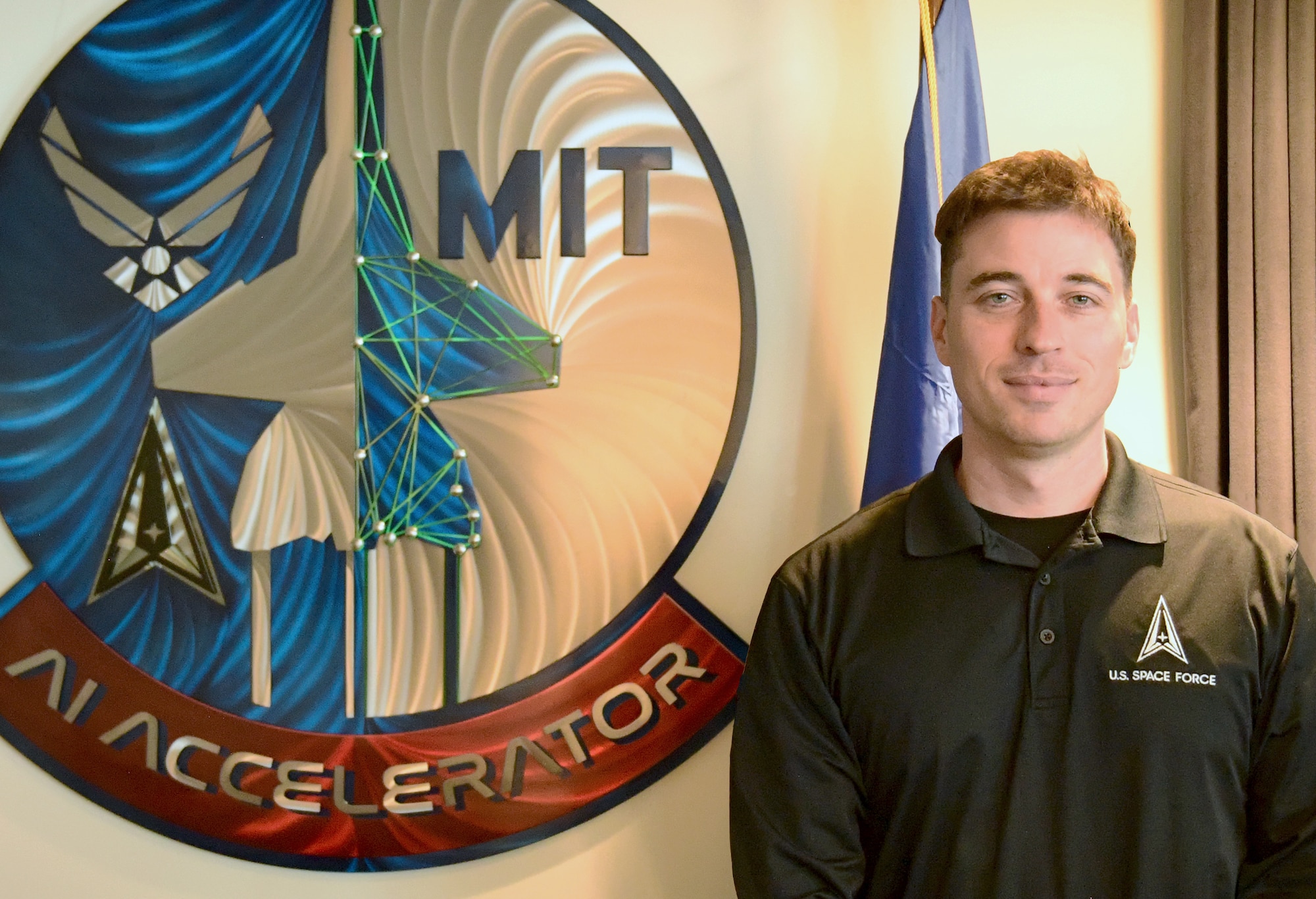 Space Force Guardian, Master Sgt. Jerry Brock, operations flight chief with the 4th Space Operations Squadron stands alongside the DAF-MIT AI Accelerator wall mount during a trip to Cambridge, Mass., March 16, 2026.