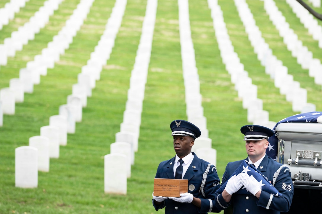 Two service members in ceremonial dress, one holding a box, the other a folded American flag, walk in front of a casket in a cemetery.
