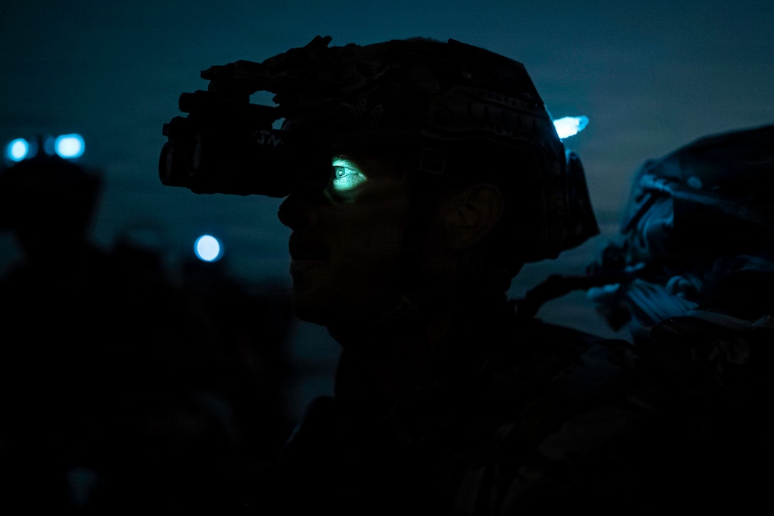 A close-up of a sailor using night vision binoculars in the dark as a light shines on the sailor's eye.