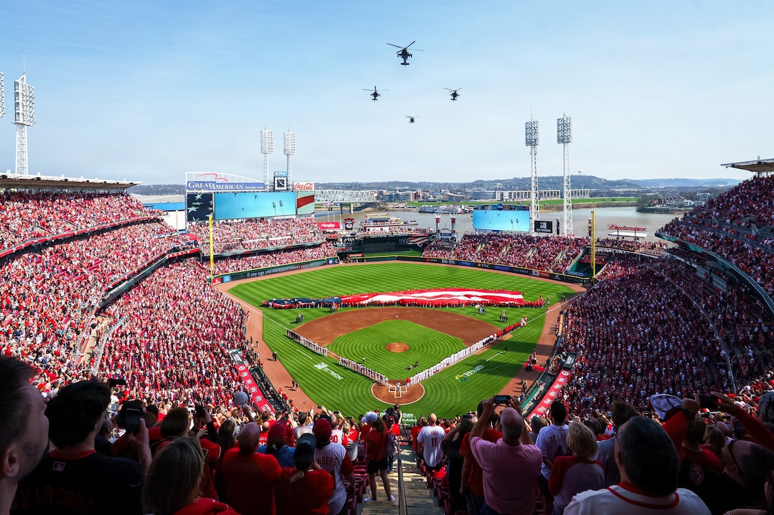 Four helicopters fly in a diamond formation in a blue sky over a baseball field as thousands of fans in red watch from the stands.