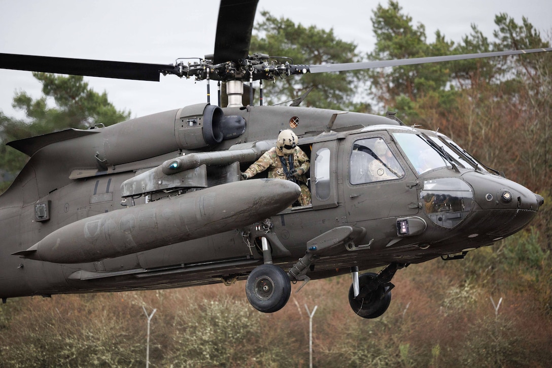A soldier leans out the open doorway of a helicopter as it flies in front of a wooded area on a gloomy day.