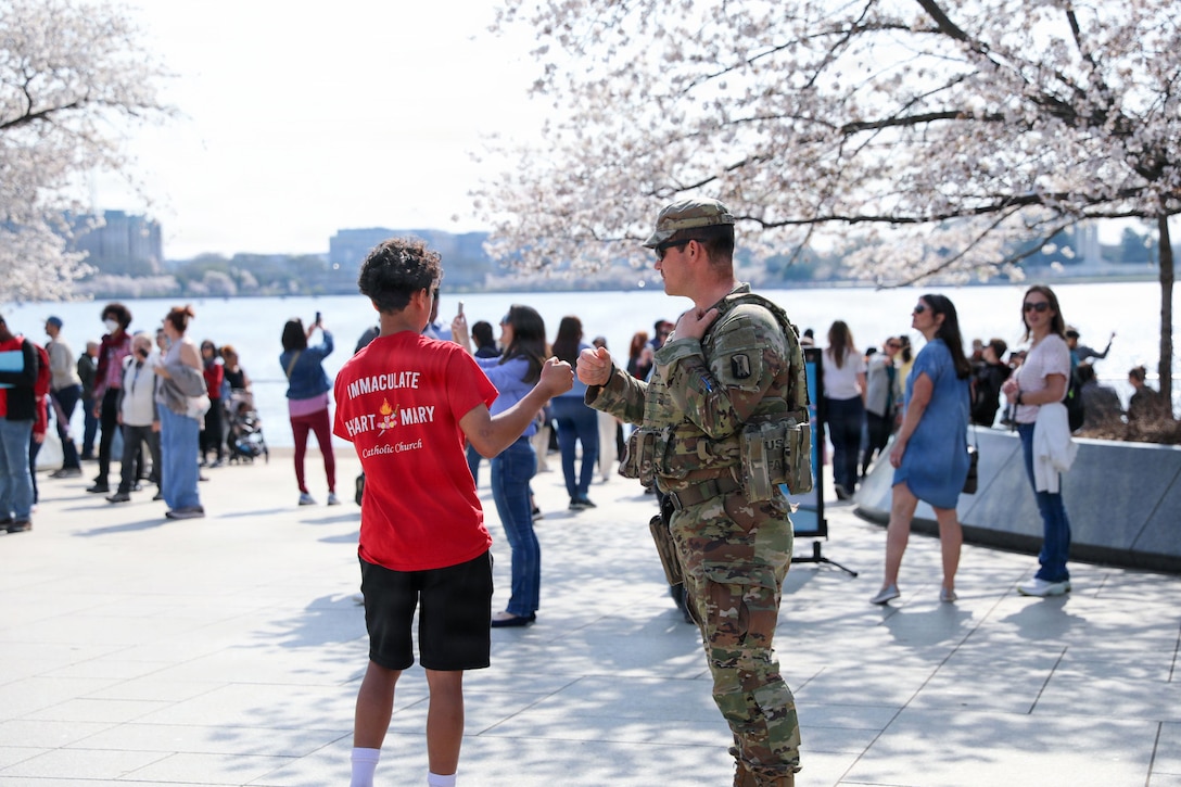 A soldier fist-bumps a young tourist as fellow tourists gather and take pictures of cherry blossom trees in front of a body of water.
