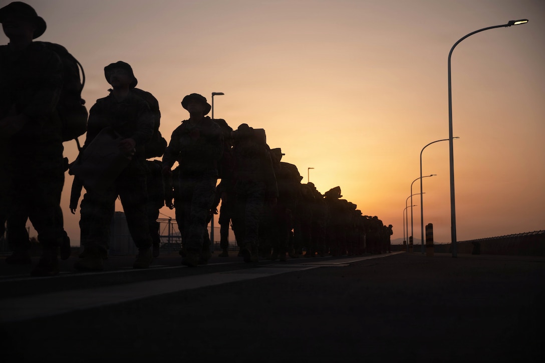 Marines and sailors in tactical gear hike to the left in silhouette under an orangish sky.