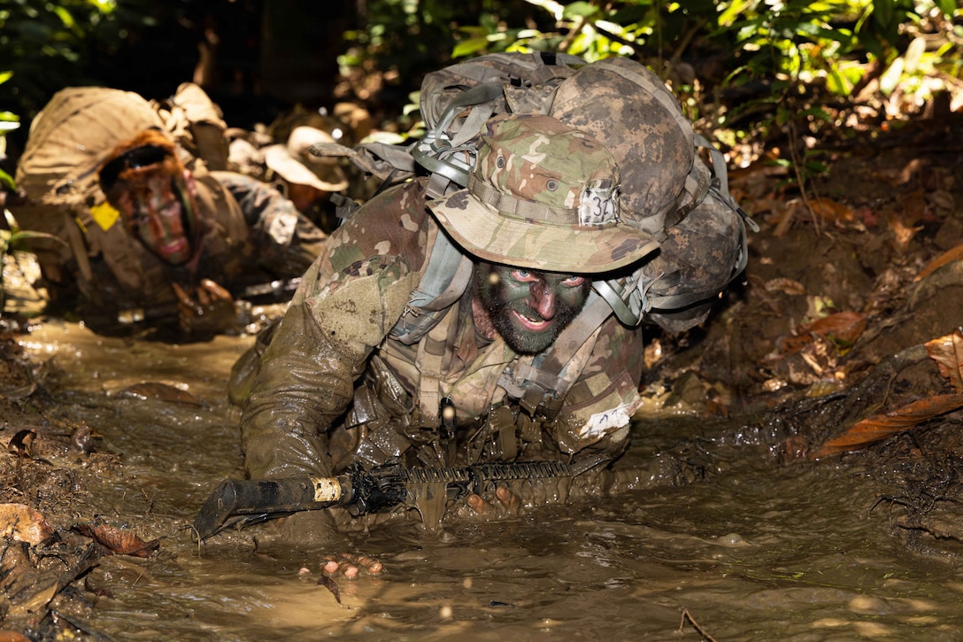 Soldiers wearing camouflage and tactical gear low crawl through the mud with weapons in a wooded area.