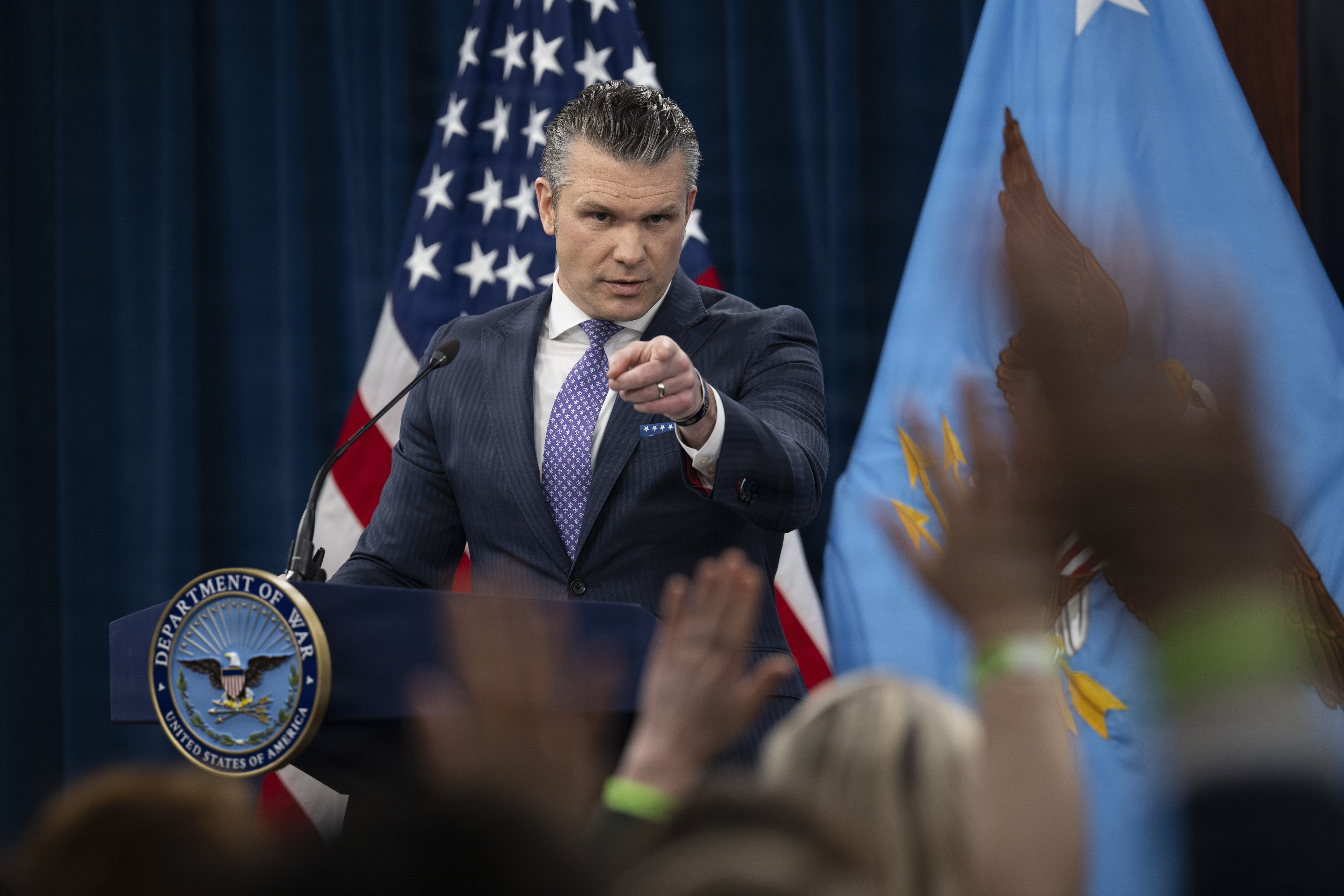 A person in a business suit standing behind a lectern points toward people sitting in front of him, some with their hands raised; an American flag and a blue flag are in the background.