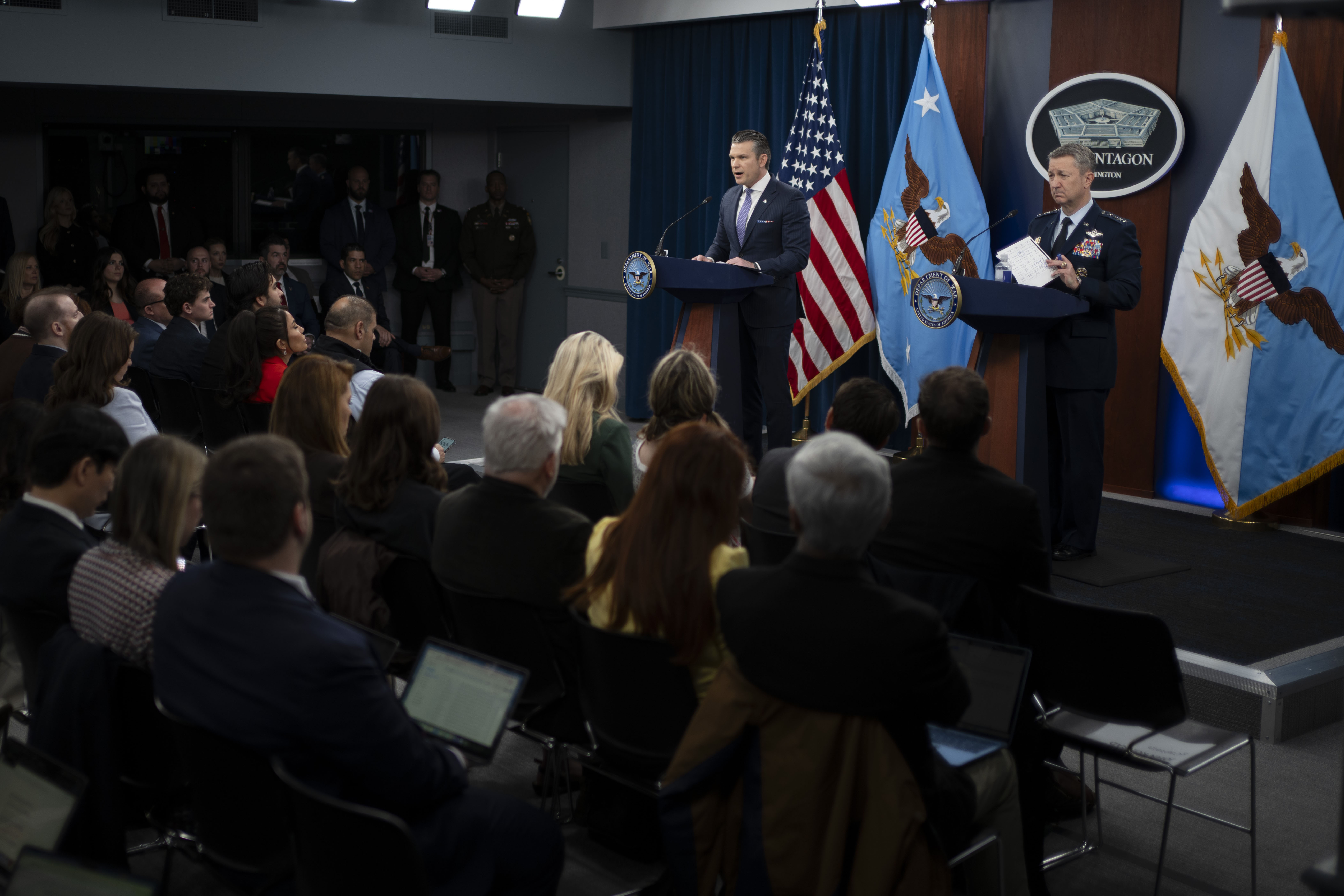 A person in a business suit and a person in a military dress uniform stand behind lecterns indoors and address people sitting in front of them; three flags are in the background.