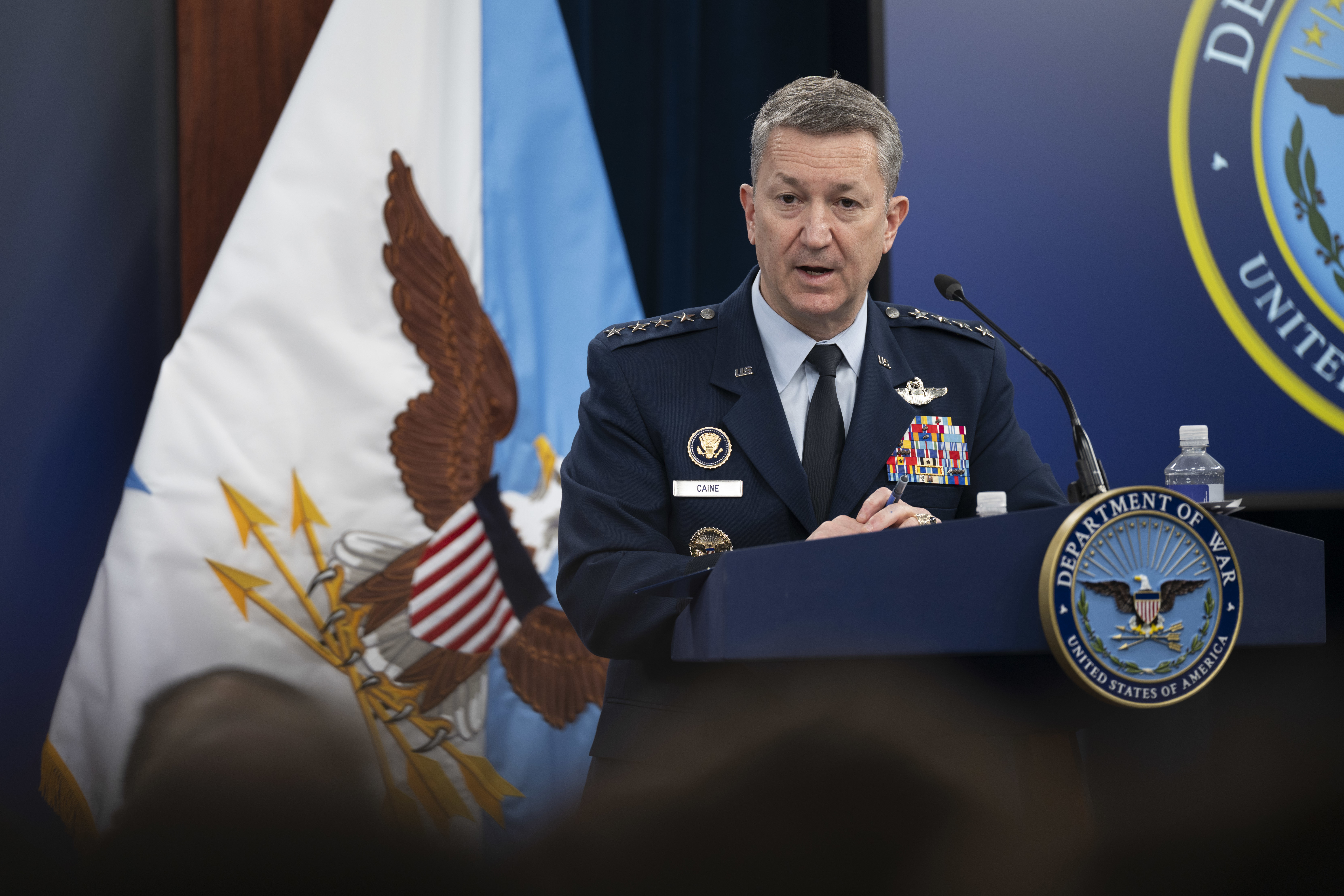 A person in a formal military uniform standing behind a lectern addresses people sitting in front of him; a blue and white flag with an eagle on it is in the background. 