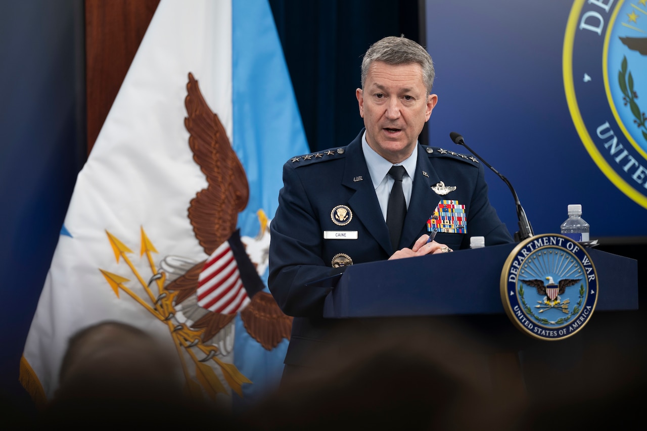 A person in a formal military uniform standing behind a lectern addresses people sitting in front of him; a blue and white flag with an eagle on it is in the background.