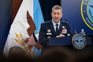 A person in a formal military uniform standing behind a lectern addresses people sitting in front of him; a blue and white flag with an eagle on it is in the background.