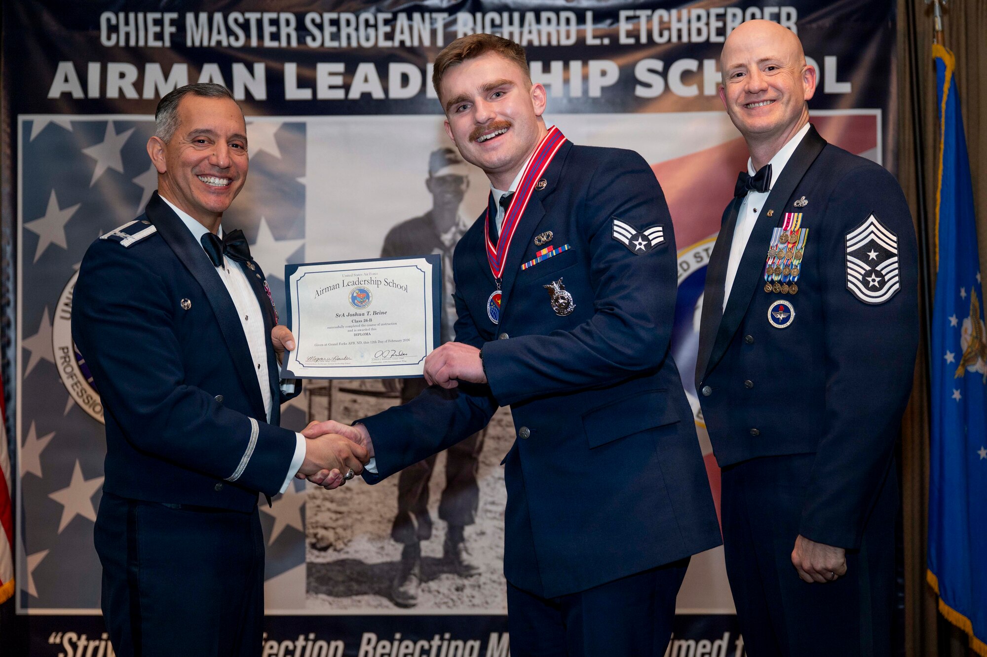 Col. Alfred Rosales, 319th Reconnaissance Wing commander, and Chief Master Sgt. William Gruenes, 319th RW command chief, presents Senior Airman Joshua Beine with an Airman Leadership School graduation certificate at Grand Forks Air Force Base, N.D., Feb. 12, 2026.