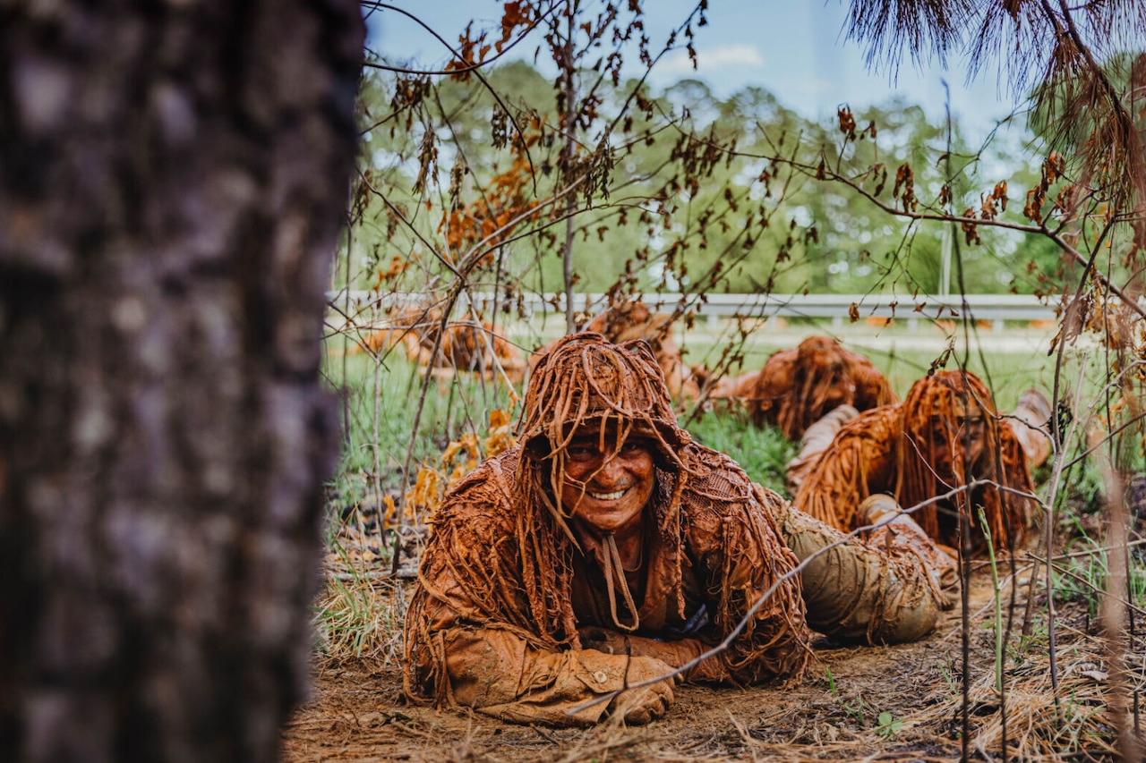 A man wearing a helmet and a ghillie suit is covered in sand, water and mud while crawling on the ground in a wooded area. There is a single line of people in similar attire crawling behind him.