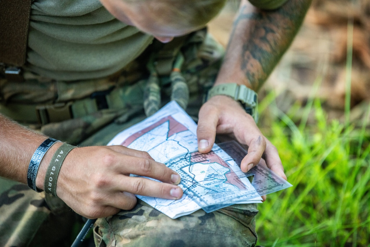 A man wearing a green T-shirt and camouflage pants kneels in a wooded area and uses a measuring device on a map.