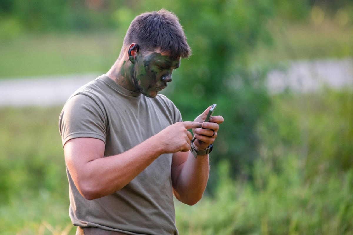A man wearing a green T-shirt and camouflage pants applies camouflage paint to his face.
