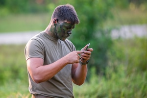 A man wearing a green T-shirt and camouflage pants applies camouflage paint to his face.