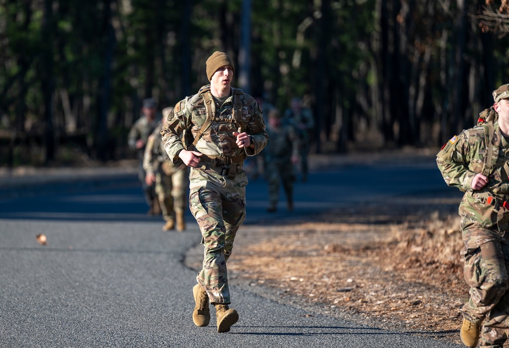 A uniformed service member jogs along a road carrying a large rucksack.