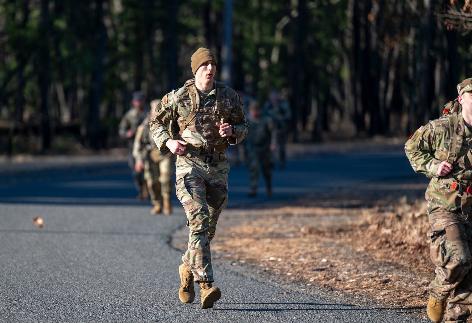 A uniformed service member jogs along a road carrying a large rucksack.