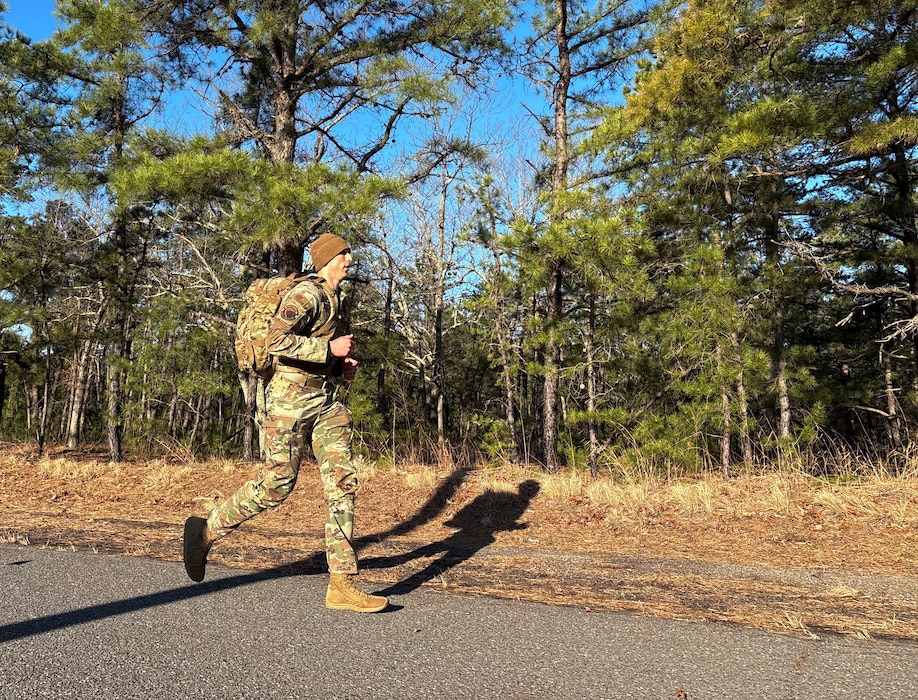 A uniformed service member jogs along a road carrying a large rucksack.