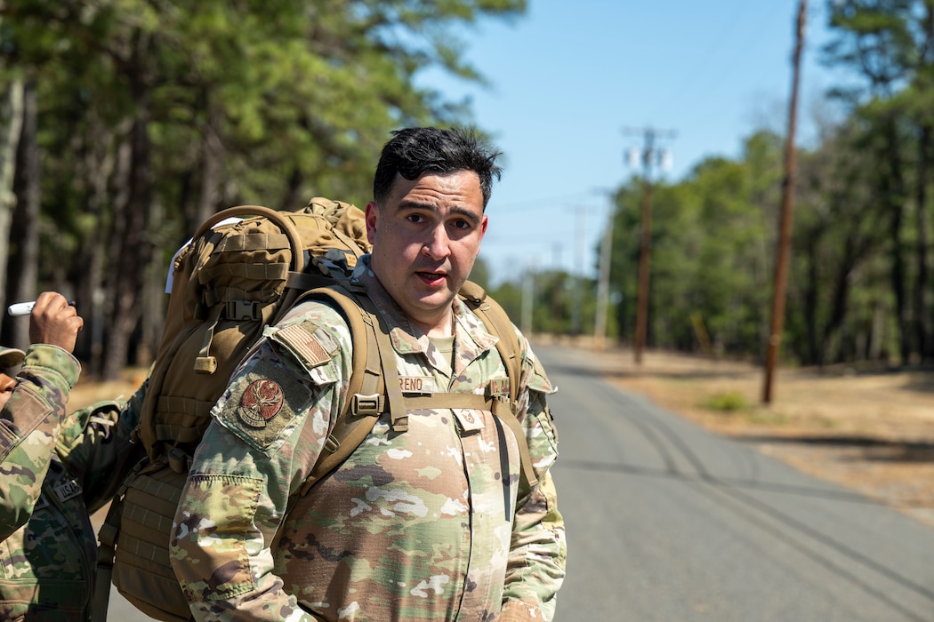A uniformed service member jogs along a road carrying a large rucksack.