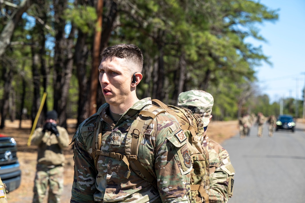 A uniformed service member jogs along a road carrying a large rucksack.