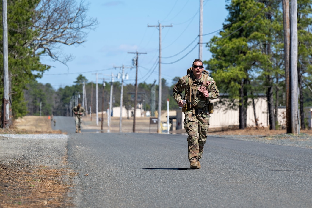 A uniformed service member jogs along a road carrying a large rucksack.