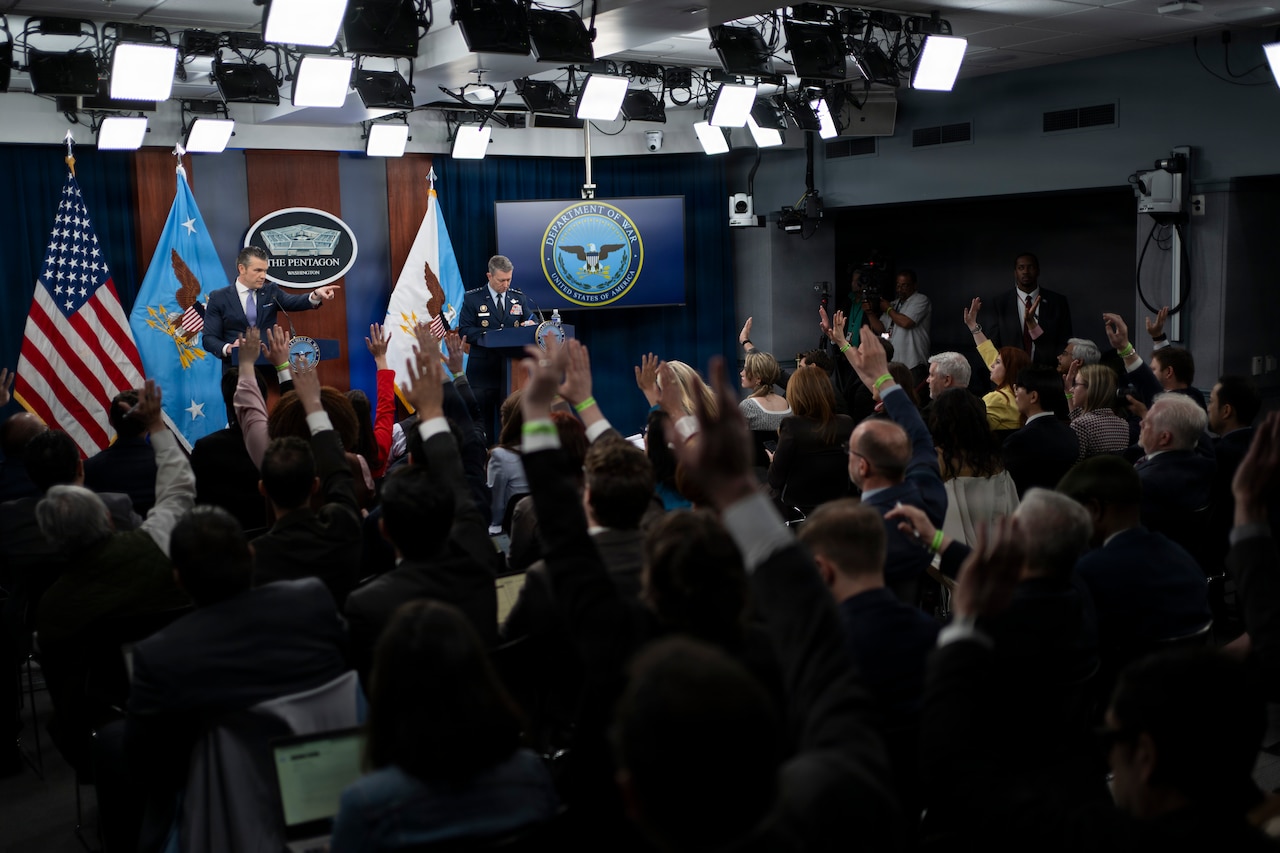 Two men stand behind lecterns speaking to an audience. In front of them are dozens of individuals, many with their hands raised.