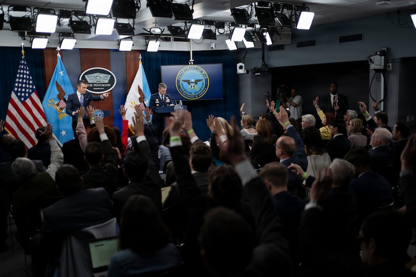 Two men stand behind lecterns speaking to an audience. In front of them are dozens of individuals, many with their hands raised.