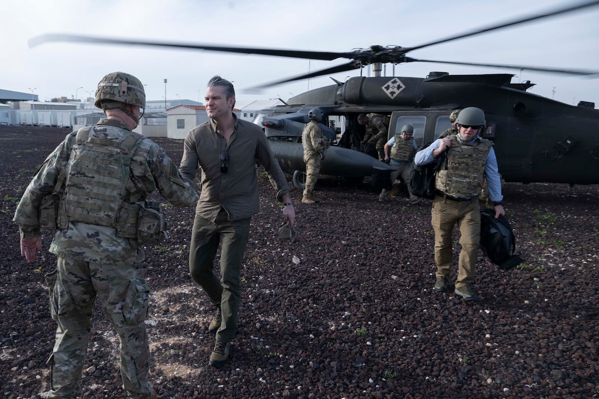 A man in civilian clothing shakes hands with a person in a camouflage military uniform; behind them, other individuals exit a helicopter.