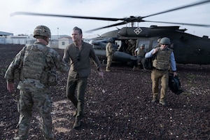 A man in civilian clothing shakes hands with a person in a camouflage military uniform; behind them, other individuals exit a helicopter.